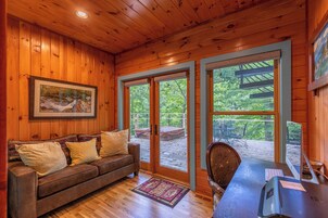 A wooden cabin interior with a wooden ceiling, a sofa with cushions, a desk with a computer, and large windows offering a view of a deck and greenery outside.