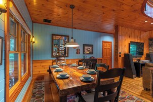Una mesa de comedor de madera dispuesta con platos y vasos, rodeada de sillas, en una habitación con paredes de madera y un ventanal grande.