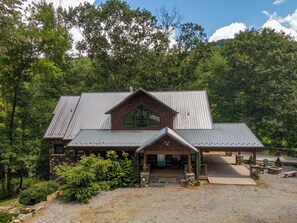 A rustic cabin with a stone facade, a large window, and a metal roof, surrounded by dense forest.