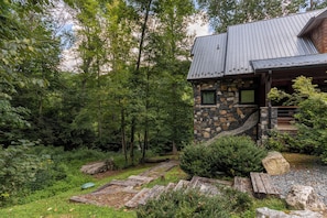 A rustic stone house with a metal roof, surrounded by dense forest and a stone pathway leading to the entrance.