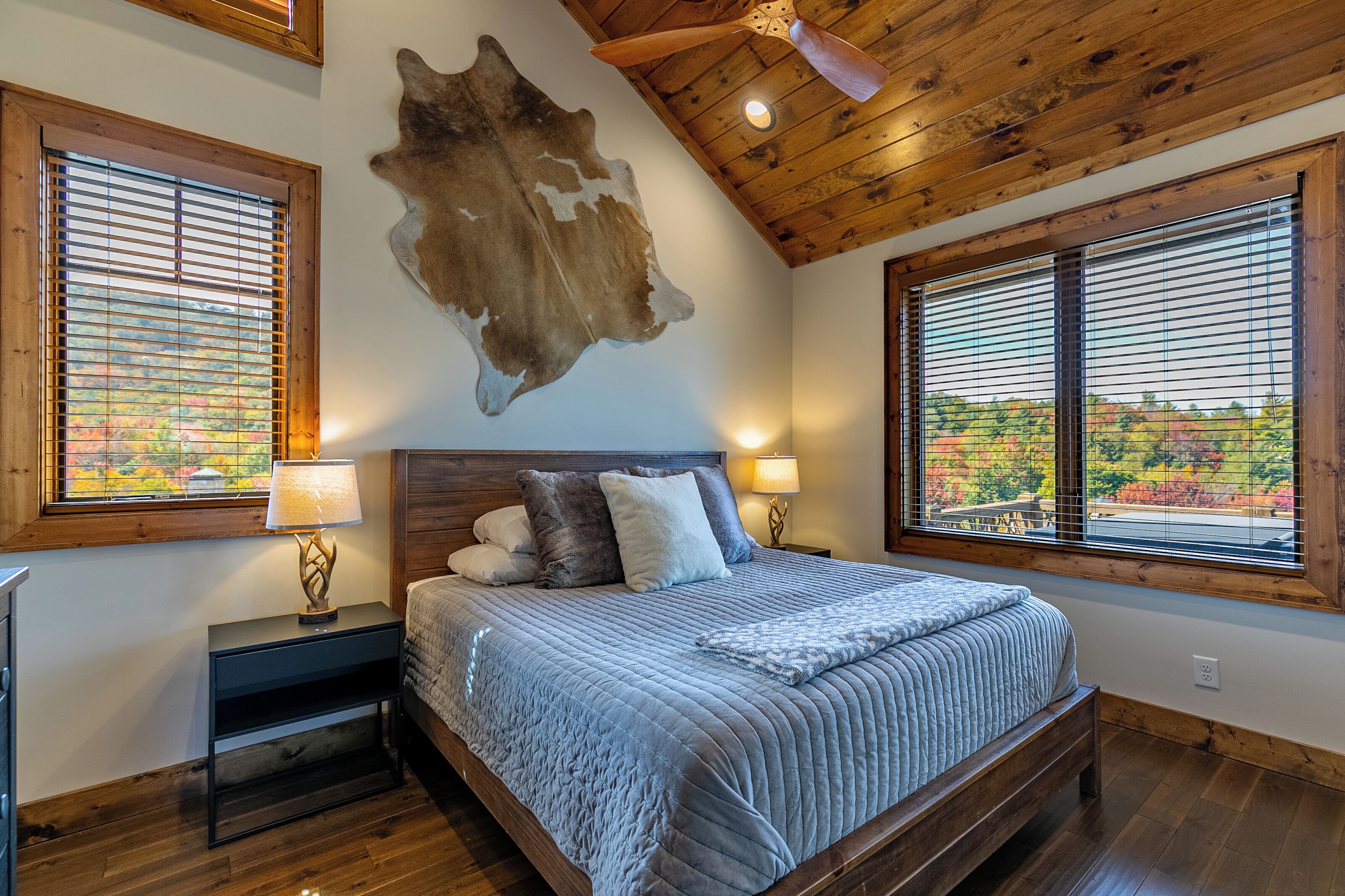 A bedroom with a wooden ceiling, a large bed with a blue quilt, two bedside lamps, and a view of autumn foliage through the windows.