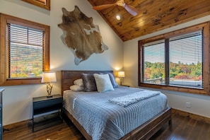 A bedroom with a wooden ceiling, a large bed with a blue quilt, two bedside lamps, and a view of autumn foliage through the windows. - Cabearnet at Eagles Nest (Banner Elk)