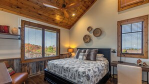 Ein Schlafzimmer mit Holzboden, einem Bett mit gemustertem Überwurf, zwei Fenstern mit Jalousien, einem Nachttisch mit Lampe und einem Schreibtisch mit Stuhl.
