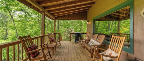 A wooden deck with rocking chairs and a grill, surrounded by trees.