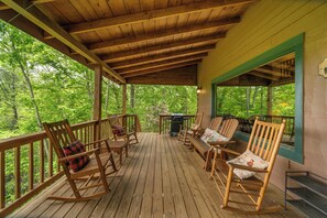 Eine Holzterrasse mit Schaukelstühlen und Grill, umgeben von Bäumen.