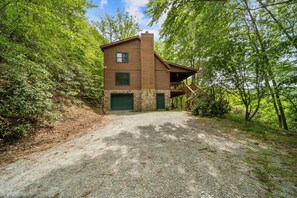 Eine zweistöckige Blockhütte mit Steinunterbau, einer Veranda und einer Schotterzufahrt, umgeben von dichtem Wald.