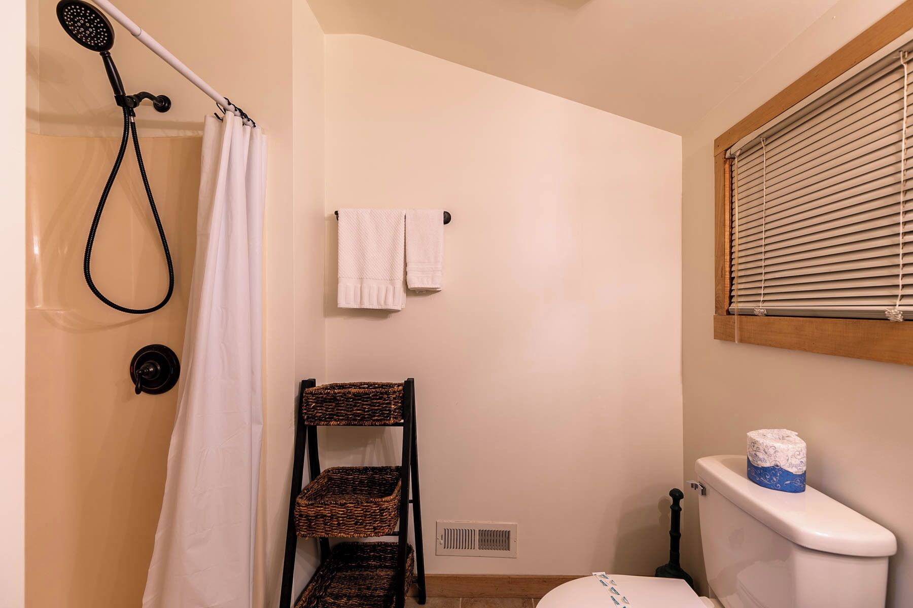 A bathroom with a shower, a toilet, a window with blinds, and a towel rack.