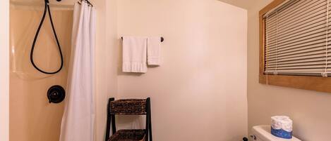 A bathroom with a shower, a toilet, a window with blinds, and a towel rack.