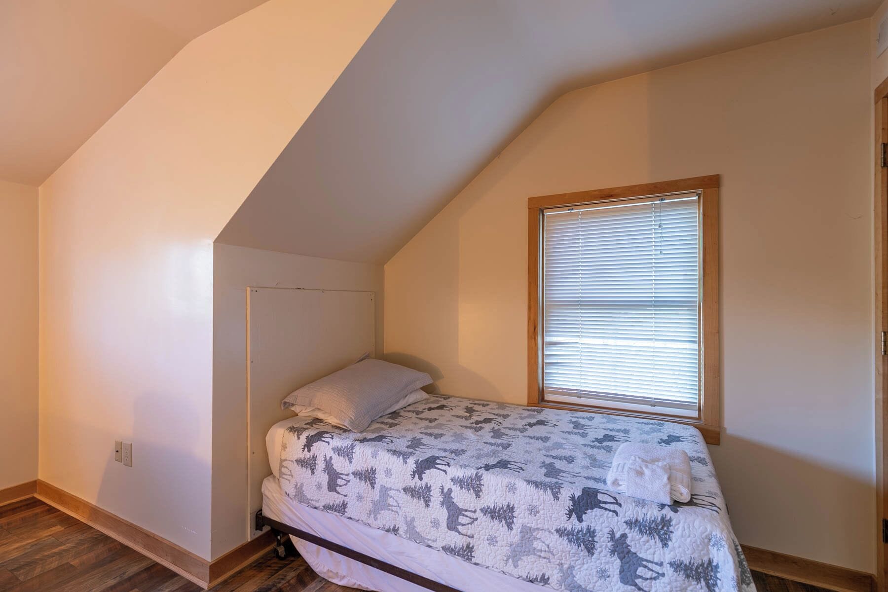 A small bedroom with a sloped ceiling, a single bed with a patterned bedspread, a window with blinds, and wooden flooring.