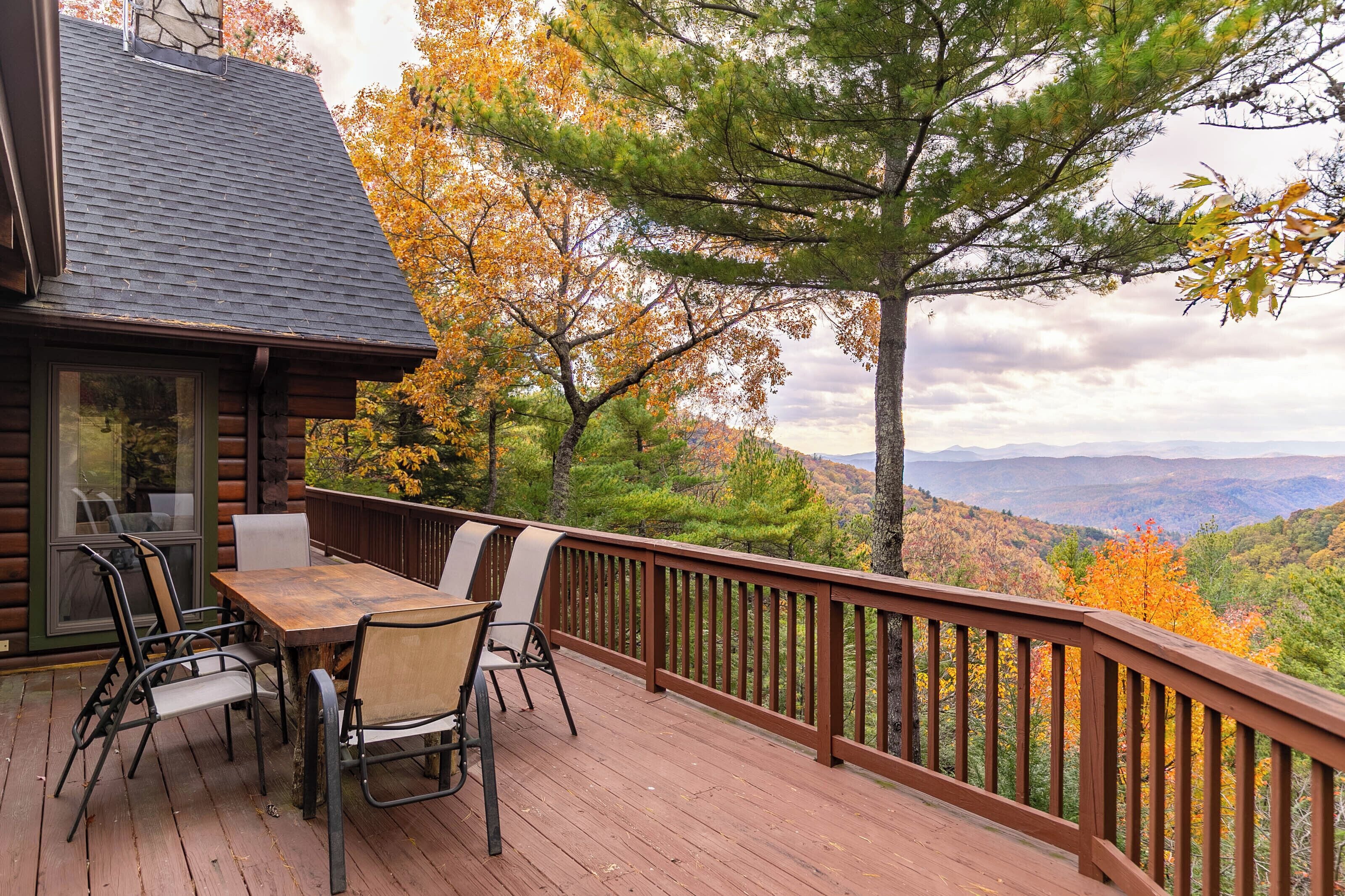 A wooden deck with a table and chairs, overlooking a forested landscape with autumn foliage.