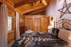 Un dormitorio tipo cabaña de troncos con una cama, un aparador, una ventana con cortinas y techo de madera.