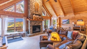 A log cabin interior with a stone fireplace, leather furniture, and a view of autumn foliage. - Angels Envy (Lenoir)