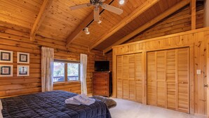 A wooden cabin bedroom with a bed, a ceiling fan, a window with curtains, a dresser, and a set of wooden closets. - Angels Envy (Lenoir)