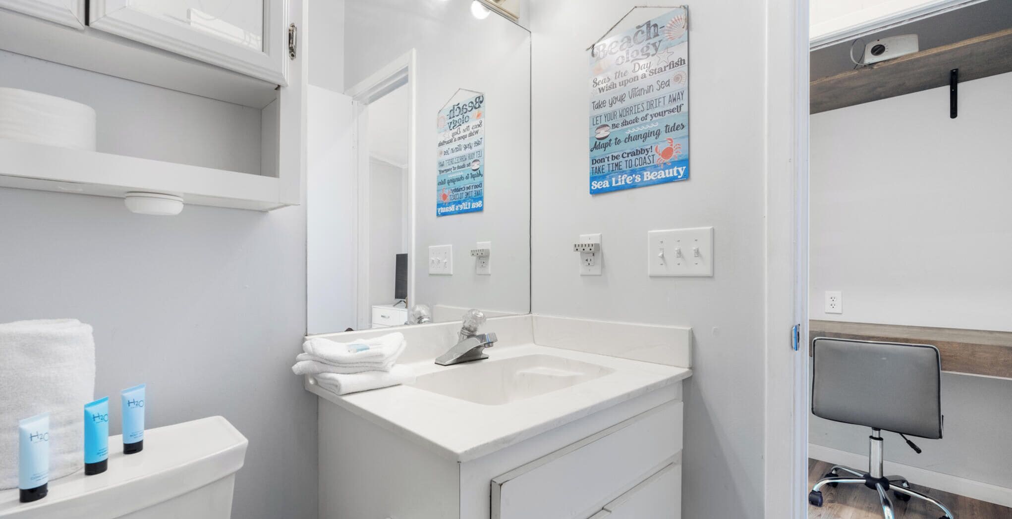 A modern bathroom with a white sink, mirror, and shelves. There are toiletries on the shelf and a chair in the corner.