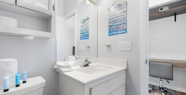 A modern bathroom with a white sink, mirror, and shelves. There are toiletries on the shelf and a chair in the corner. - Pinetree Parlor (Panama City Beach)