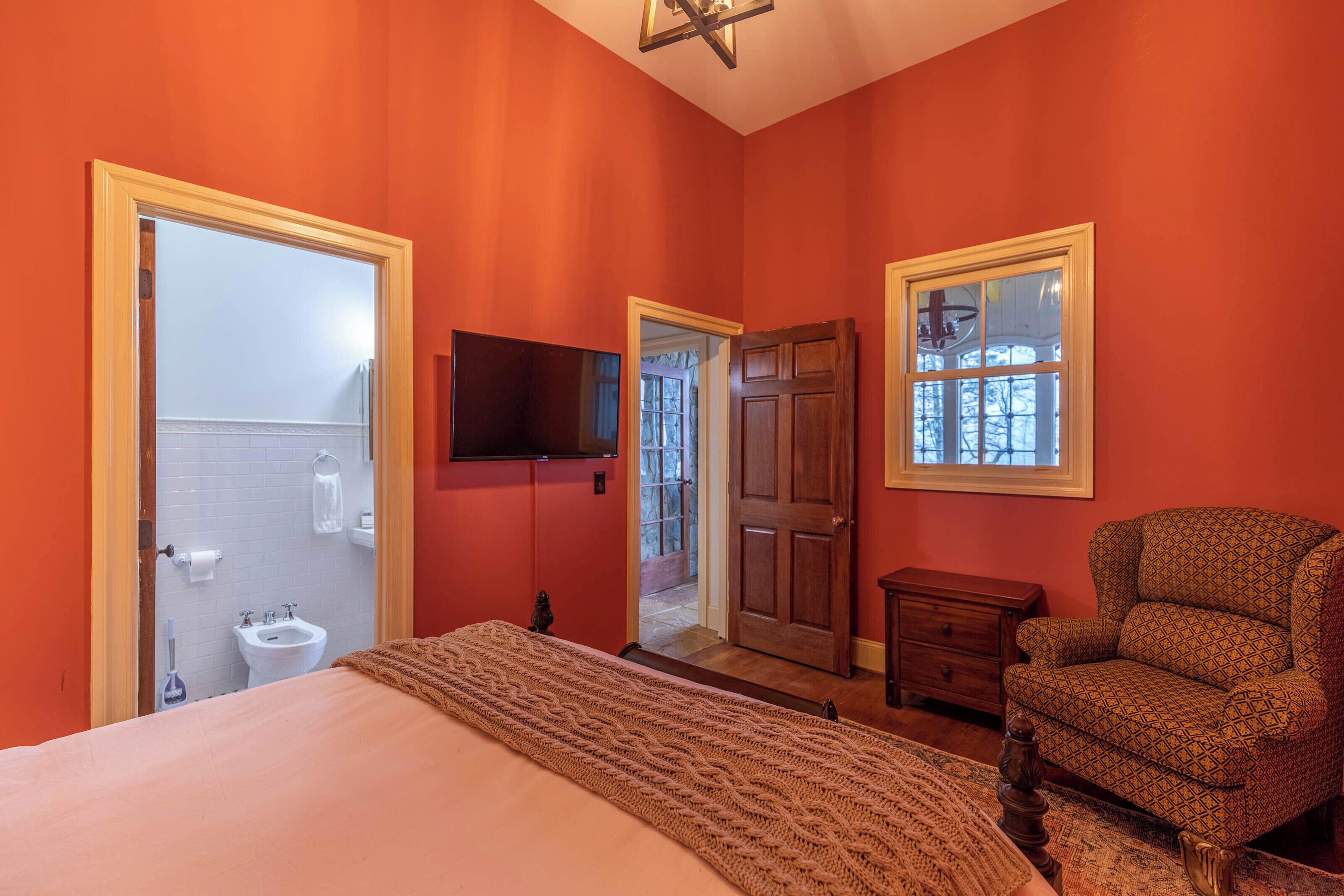 A bedroom with a large bed, a wooden chair, a television, and a door leading to a bathroom.