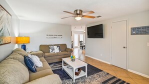 Un salon avec un ventilateur de plafond, un canapé, une table basse et un téléviseur fixé au mur.