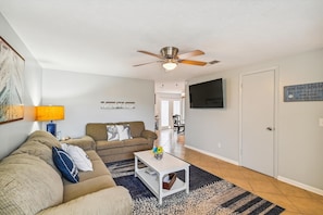 Ein Wohnzimmer mit Deckenventilator, einer Couch, einem Couchtisch und einem Fernseher an der Wand.