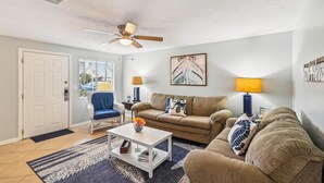 Un salon avec un ventilateur de plafond, un canapé, une table basse et un fauteuil. Il y a une fenêtre avec des stores, une porte et une lampe fixée au mur.