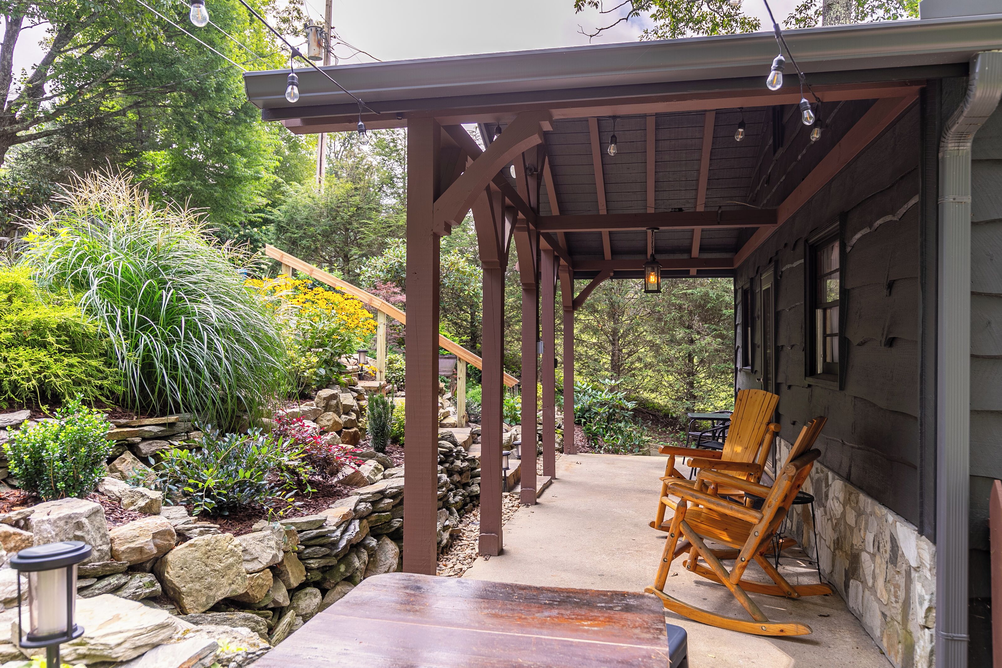 A wooden porch with a covered seating area, stone walls, and a variety of plants.