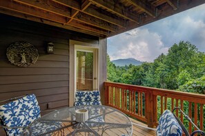 Une terrasse en bois avec une table ronde et des chaises, une porte et une vue sur une chaîne de montagnes boisée.