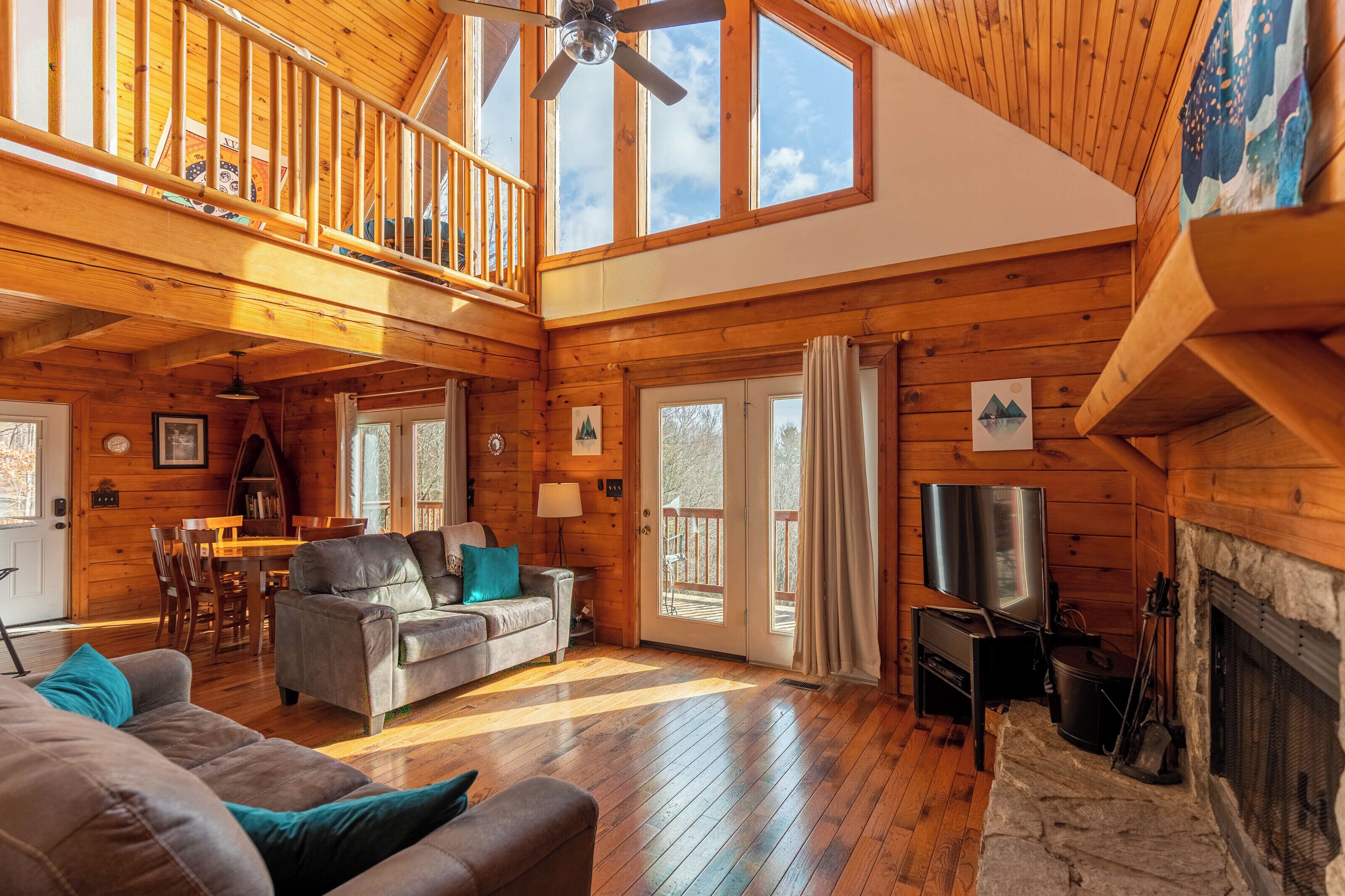 A cozy log cabin interior with a loft bedroom, a fireplace, a dining area, and a living room with a sofa and TV.