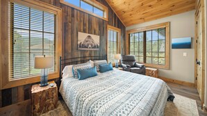A bedroom with a wooden ceiling, large windows, a bed with patterned bedding, a chair, and a nightstand with a lamp. - River Rock (Banner Elk)