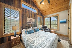 Ein Schlafzimmer mit Holzbalkendecke, großen Fenstern, einem Bett mit gemusterten Betttextilien, einem Sessel und einem Nachttisch mit Lampe.
