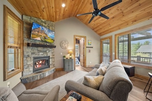 A living room with a stone fireplace, a ceiling fan, and a large window. - River Rock (Banner Elk)