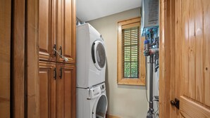 A laundry room with a stacked washer and dryer, wooden cabinets, a window with blinds, and a vent system. - River Rock (Banner Elk)