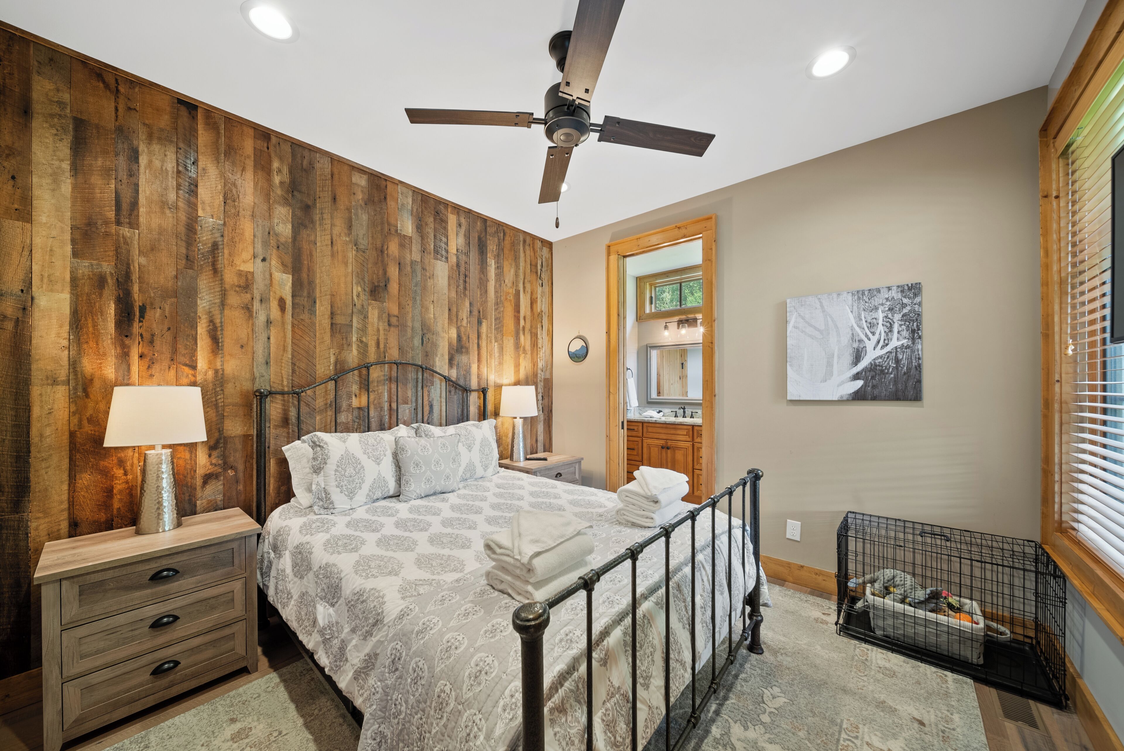A bedroom with a wooden wall, a bed with a metal frame, a ceiling fan, a nightstand, a lamp, a dog crate, and a window with blinds.