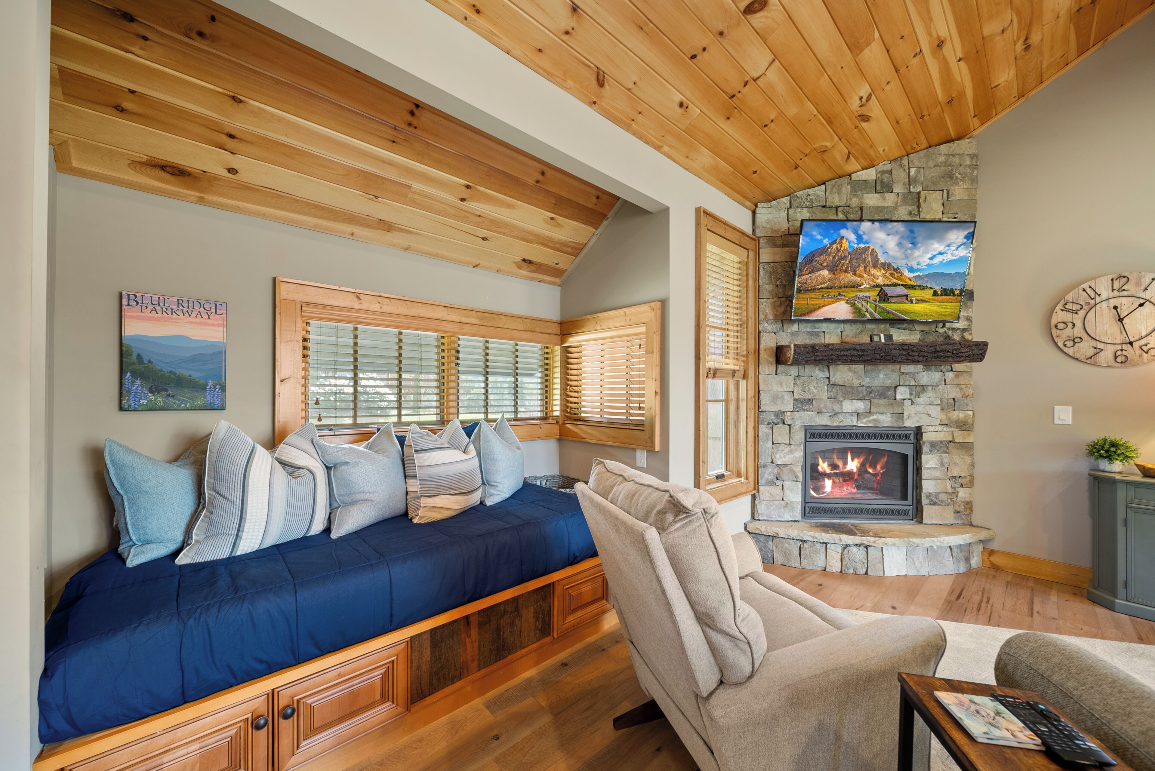 A cozy living room with a wooden ceiling, a stone fireplace, and a large window with blinds.