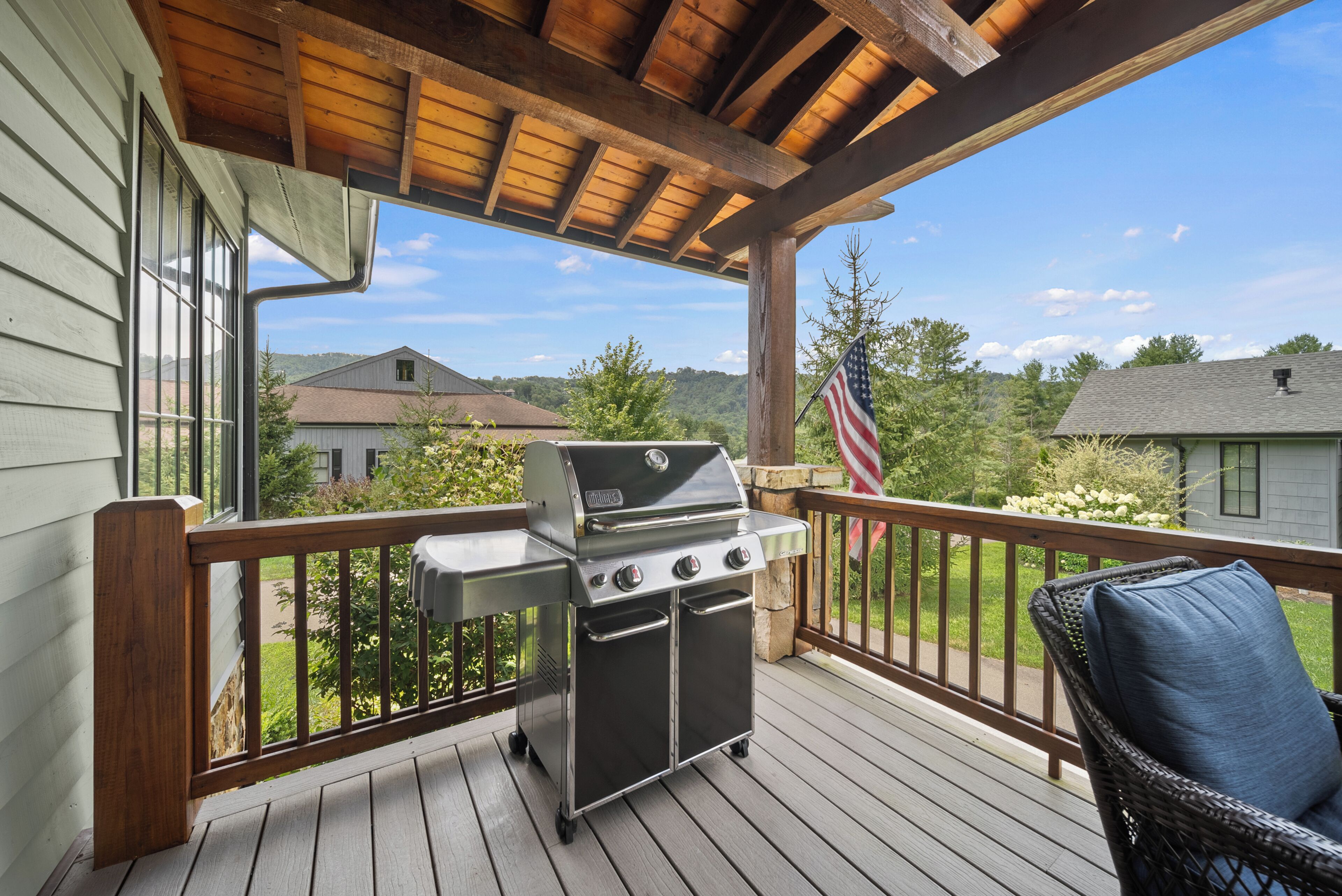 A covered deck with a grill, a wicker chair, and a view of a house and trees.