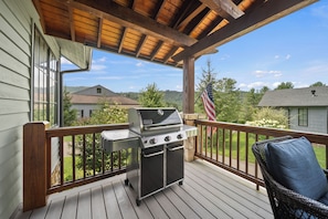 Eine überdachte Terrasse mit Grill, einem Korbstuhl und Blick auf ein Haus und Bäume.