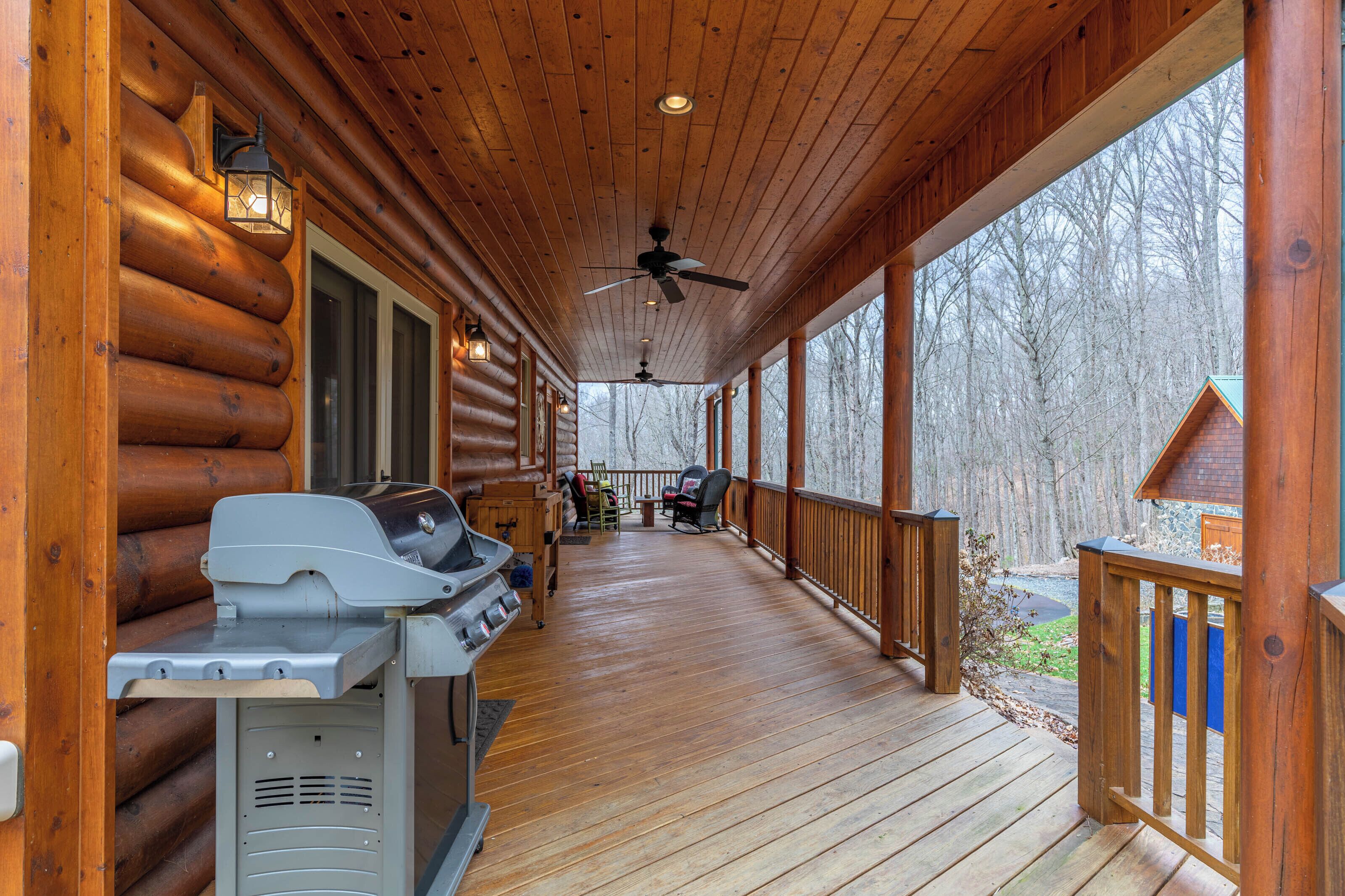A covered wooden deck with a grill, a ceiling fan, and a view of the surrounding forest.