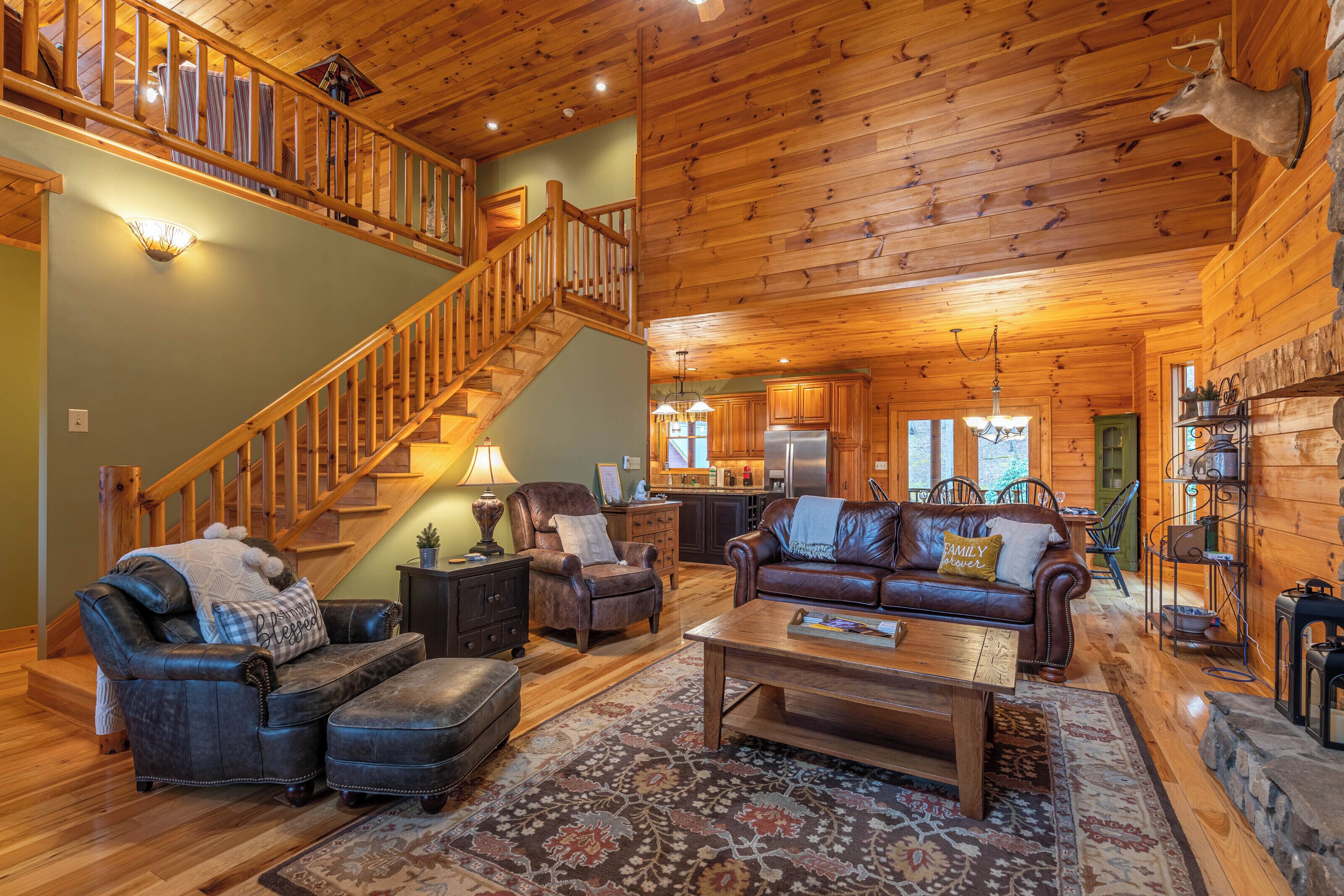 A spacious log cabin interior with a wooden staircase, leather furniture, and a stone fireplace.