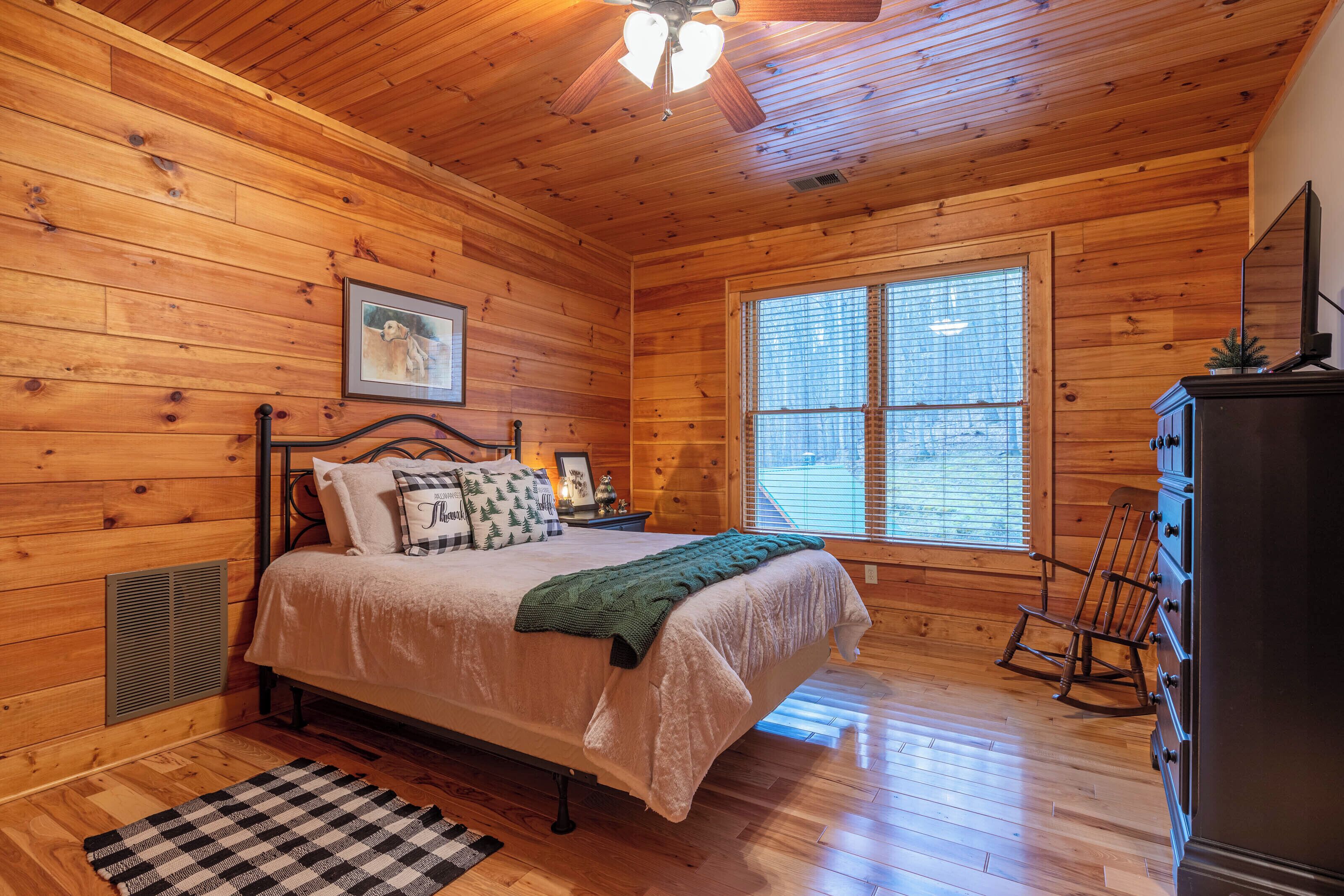 Une chambre à coucher aux murs lambrissés, avec un lit, un fauteuil à bascule, une commode et une fenêtre équipée de stores.