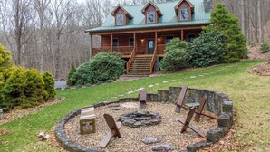 A log cabin with a stone fire pit and a wooden bench, surrounded by a stone wall and a grassy area. - The Forge (Lansing)