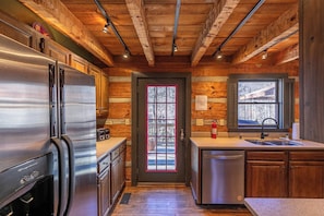 A compact kitchen with wooden walls, a stainless steel refrigerator, a dishwasher, and a window with shutters. - A River Runs Through It (Vilas)