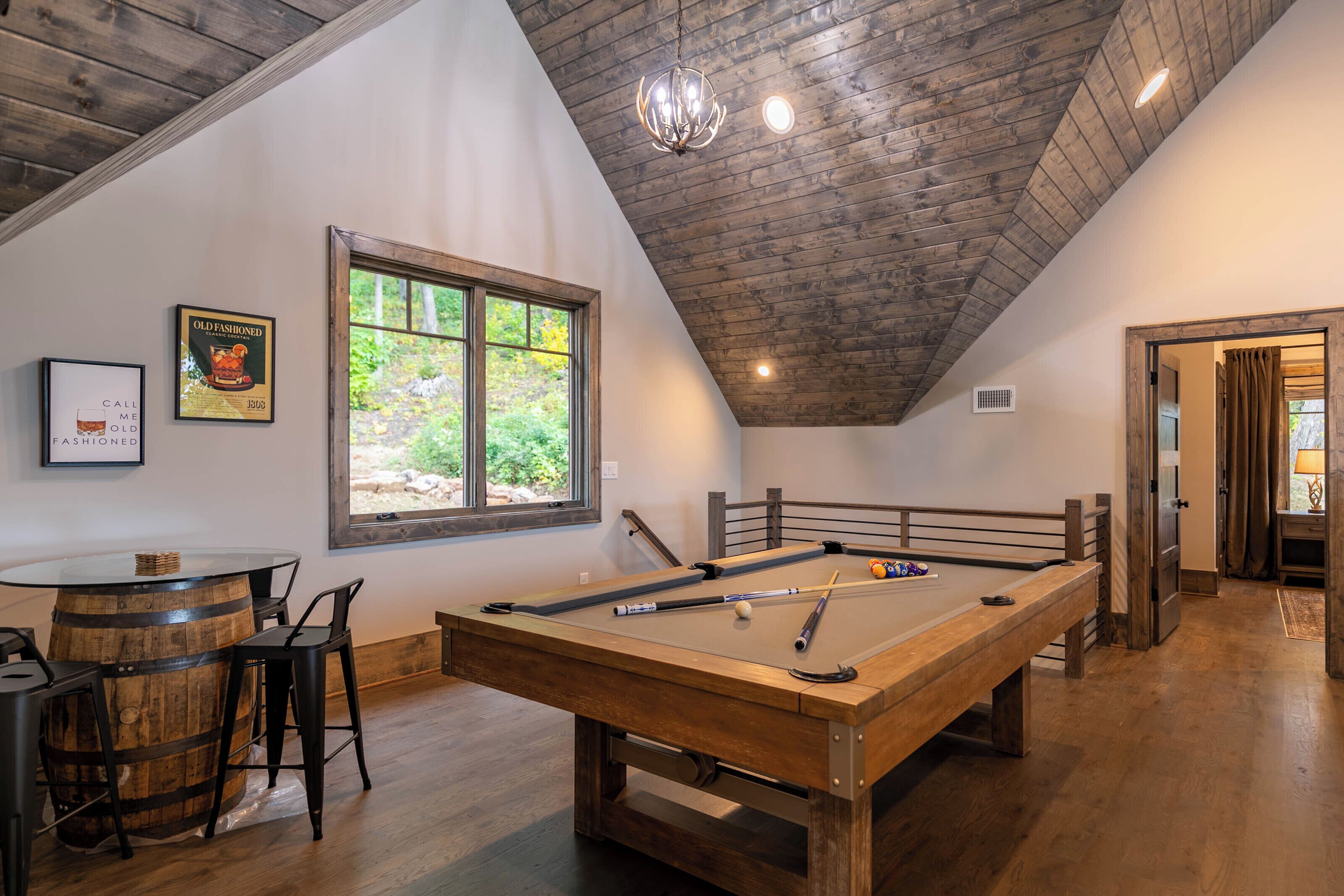 A pool table in a room with a wooden ceiling, a window with a view of greenery, and a bar area with stools.