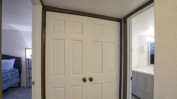 A closed white door with a dark frame, a bed with a blue blanket, and a bathroom with a white sink and cabinets. - 7035 Park City #3R (Park City)