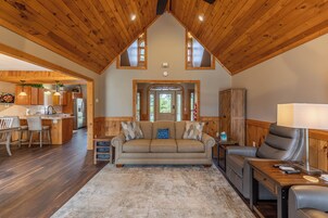 A cozy living room with a wooden ceiling, a sofa, a recliner, and a dining area with a kitchen visible in the background.