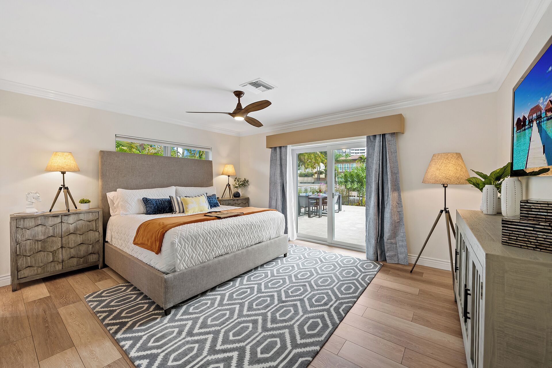 A hotel room with a bed, two bedside lamps, a ceiling fan, a TV, and a view of the outdoor area.