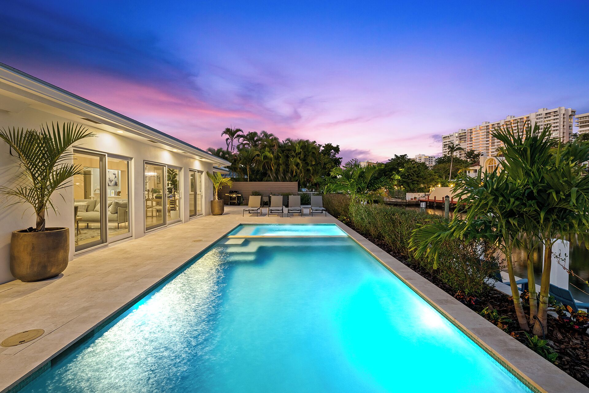 A modern swimming pool with a glass enclosure, surrounded by lush greenery and a sunset sky.