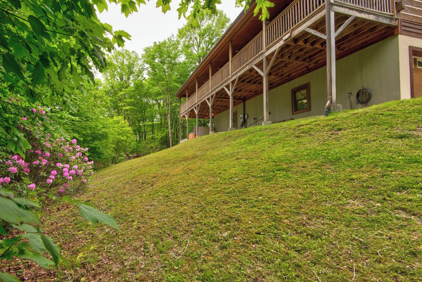 A building with a wooden deck and a covered porch, surrounded by lush greenery and a grassy slope.
