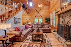 A cozy cabin interior with a wooden staircase, leather sofas, a fireplace, and a large window. - Hebron Falls Cabin (Boone)