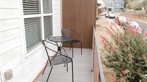 A balcony with two chairs and a table, overlooking a street with parked cars and a house in the background.