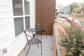 A balcony with two chairs and a table, overlooking a street with parked cars and a house in the background.