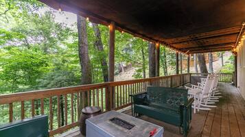 Un espacio de estar al aire libre techado con una chimenea, terraza de madera y vistas a un paisaje boscoso.