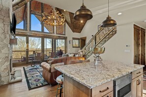 A spacious living area with a large window, a stone fireplace, a wooden staircase, and a kitchen with granite countertops. - Haven at Eagles Nest (Banner Elk)